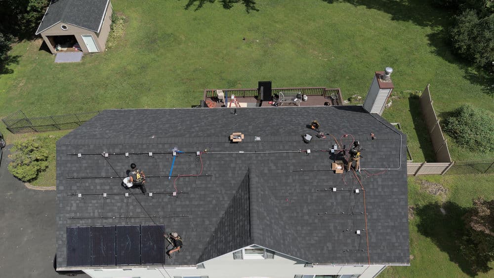 Roofing installation with workers, solar panels, and green yard area in background.